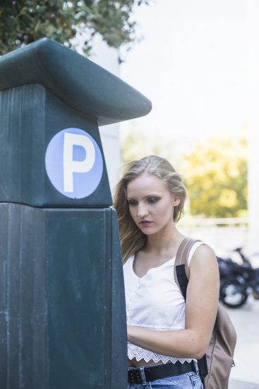 Mujer joven de pie junto a un parquímetro en una calle de la ciudad, con árboles y motocicletas de fondo.